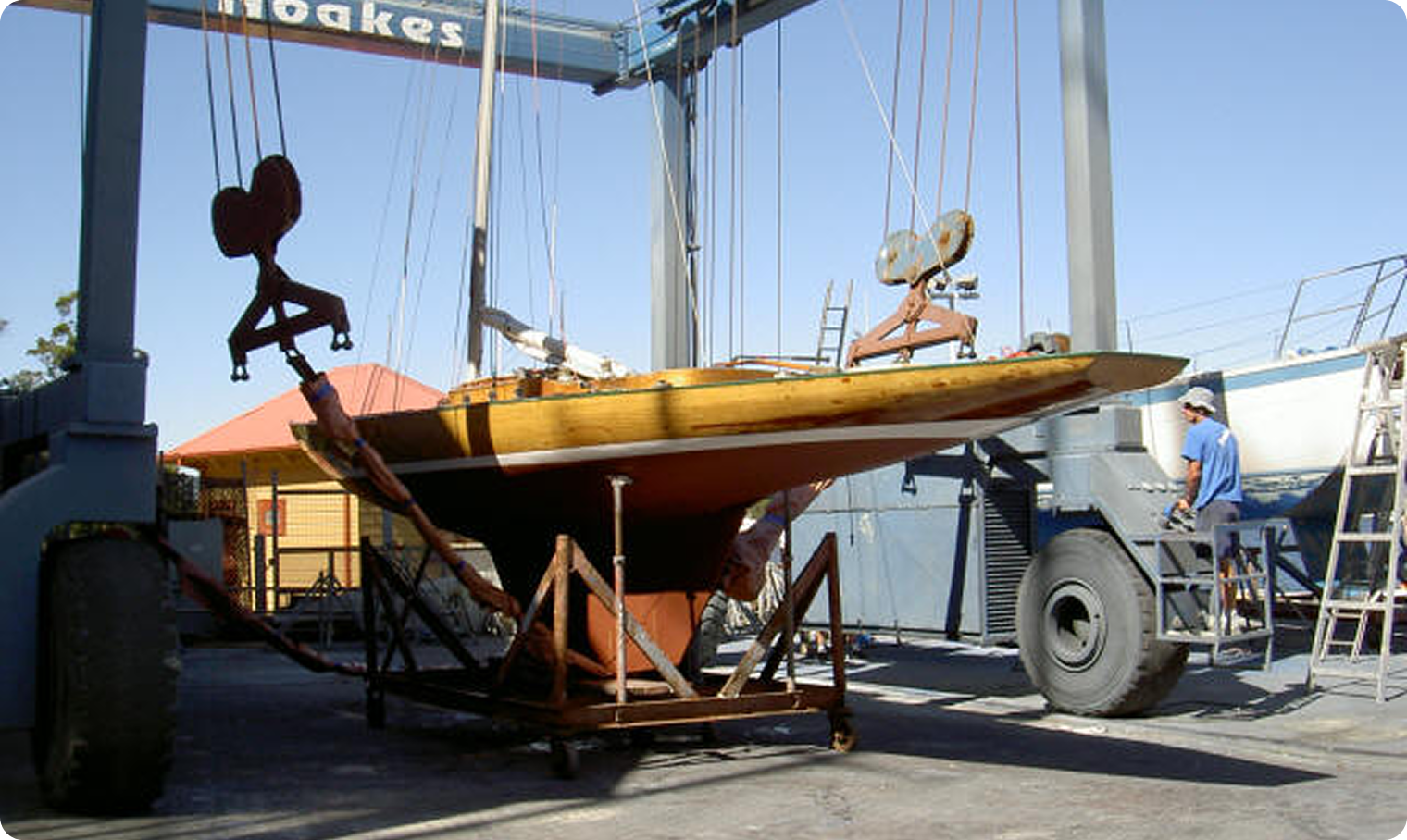 A wooden Skerry Cruiser sailing boat is being lifted by a large boat hoist at a marina. A man in a blue shirt and cap stands nearby, and another boat is partly visible on the right. The sky is clear and sunny.