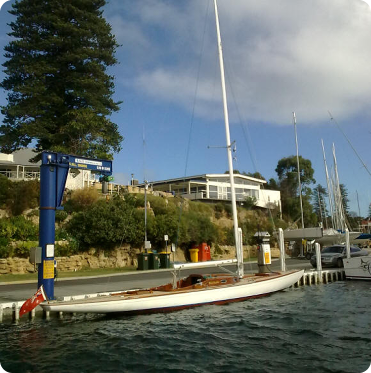 A slender white Schärenkreuzer sailboat with a tall mast is moored at a marina beside a small building, with trees and a cloudy sky in the background. The calm water reflects this classic sailing yacht among other nearby boats.