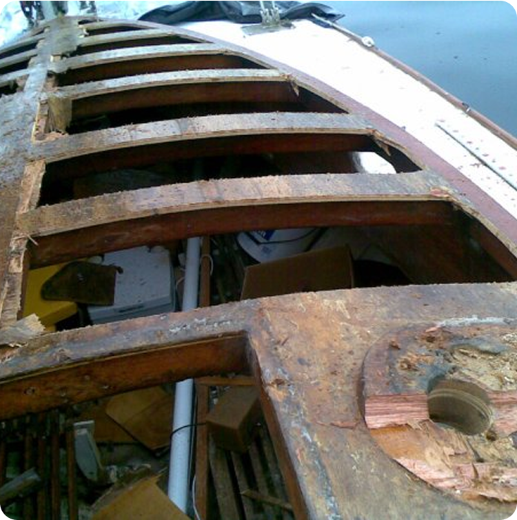 Close-up of a damaged wooden boat hull, possibly from a classic Square Metre Yacht, with several planks missing and the weathered interior structure exposed amid debris.