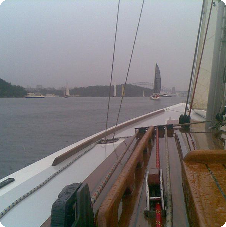 View from a sailboat deck on a cloudy day, showing the wet wooden surface, rigging, and Schärenkreuzer sailing ahead on a wide river or harbour, with land and a bridge visible in the distance.