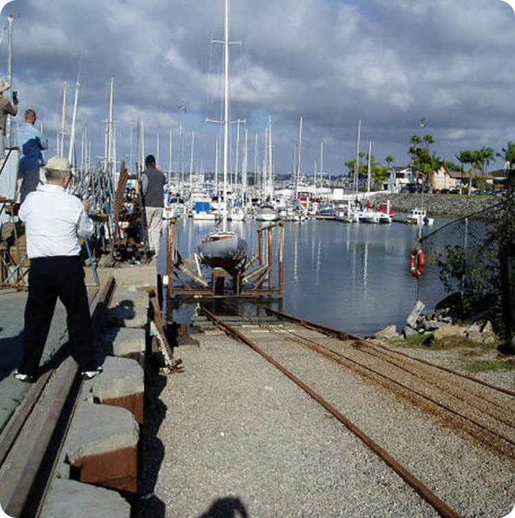 Several people stand near boat slips and tracks leading into a marina filled with sailing boats, including elegant Square Metre Yachts. The sky is partly cloudy, and palm trees are visible along the water’s edge.