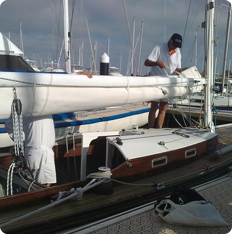 Two people in white shirts are working on a Schärenkreuzer sailboat moored at a marina, with one standing on deck and the other partially obscured. Several masts and other Classic Sailing Yachts are visible in the background.