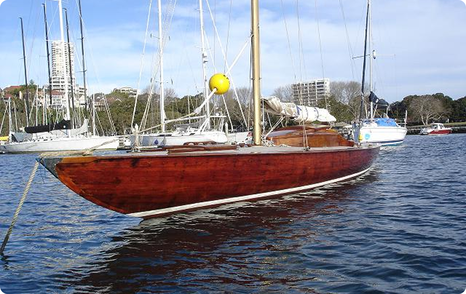 A wooden sailboat with a polished brown hull, reminiscent of classic Skerry Cruisers, is moored on calm water. White yachts and tall buildings are visible in the background under a partly cloudy sky.