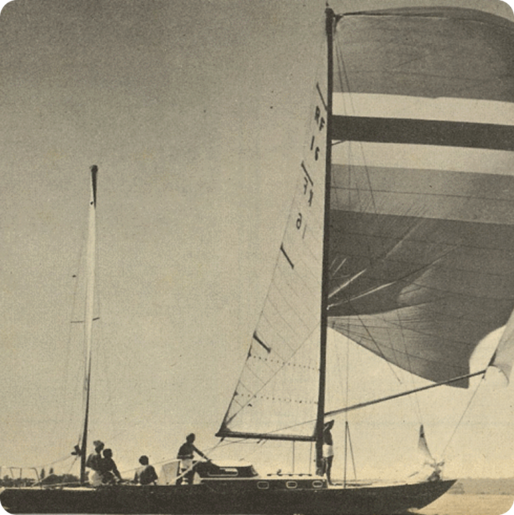 A vintage black-and-white photo of a classic sailing yacht with four people on board glides on calm water. The boat’s large, striped sail stands out as it moves steadily beneath a clear sky.