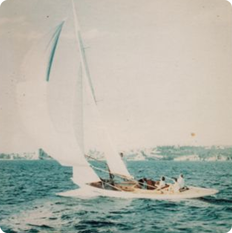 A classic sailing yacht with three people on board glides across blue water on a sunny day, with a distant shoreline and some buildings visible in the background.