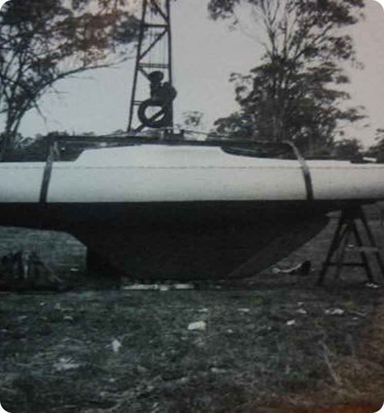 A black and white photo of a Skerry Cruiser being lifted or lowered by a crane outdoors, surrounded by trees and grass. The boat is partially elevated above the ground.