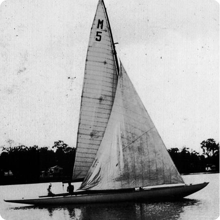 Black and white photo of a sailboat, likely a classic Schärenkreuzer or Square Metre Yacht, with the sail marked “M 5,” gliding on calm water with three people on board and a tree-lined shore in the background.