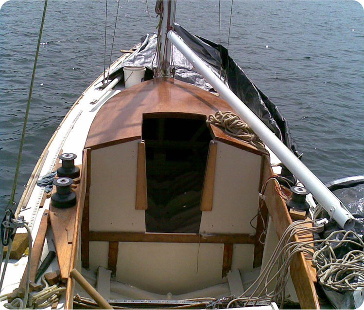 View from the deck of a Skerry Cruiser, showing the entrance to the cabin, ropes, and winches. The boat is on calm water, with the mast and partially furled sails of this elegant Square Metre Yacht visible.