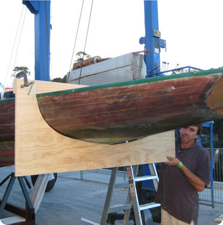 A man stands next to a wooden boat hull, possibly a Schärenkreuzer or Skerry Cruiser, held in place by a large plywood support structure, with industrial equipment and fencing visible in the background—echoing Square Metre Yachts craftsmanship.