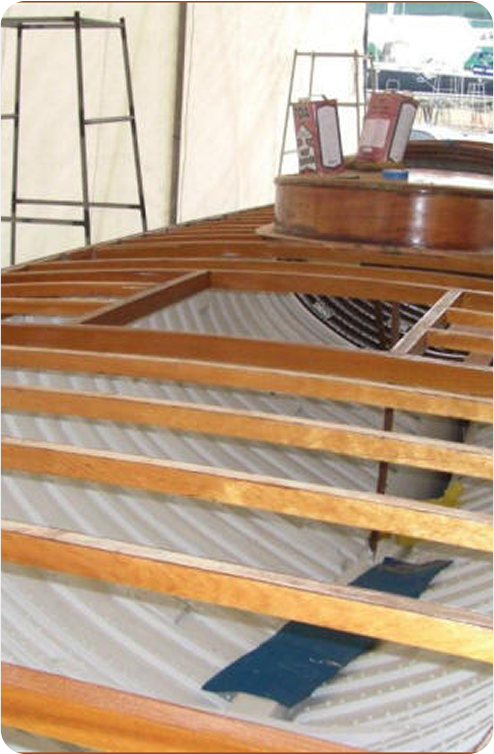 A partially constructed wooden boat, likely a classic sailing yacht or Skerry Cruiser, with visible ribs and beams, sits under a marquee. Some signs and display boards are visible in the background.