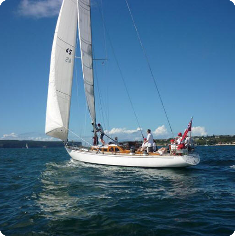 A white sailboat, reminiscent of classic Skerry Cruisers, glides on calm blue water under a clear sky, with several people on board and land and trees visible in the background.