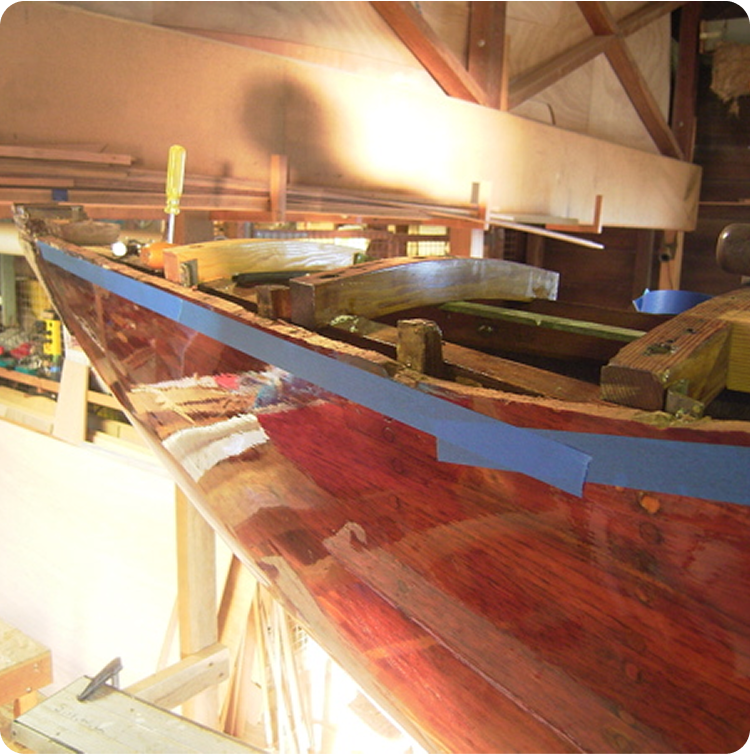 A wooden boat under construction in a workshop, with blue painter’s tape along the polished reddish-brown hull and tools resting on the unfinished wooden frame, evokes the timeless craftsmanship of Classic Sailing Yachts.