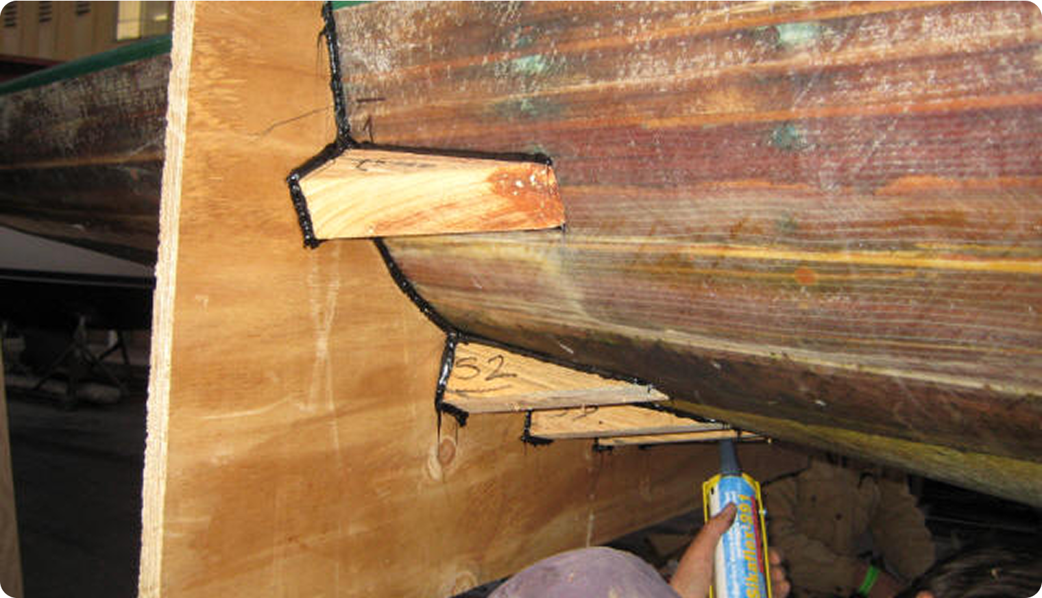 A close-up of a wooden boat hull supported by angled wooden braces, with a person applying sealant to the seams underneath—a careful restoration technique often used on Classic Sailing Yachts and Square Metre Yachts.