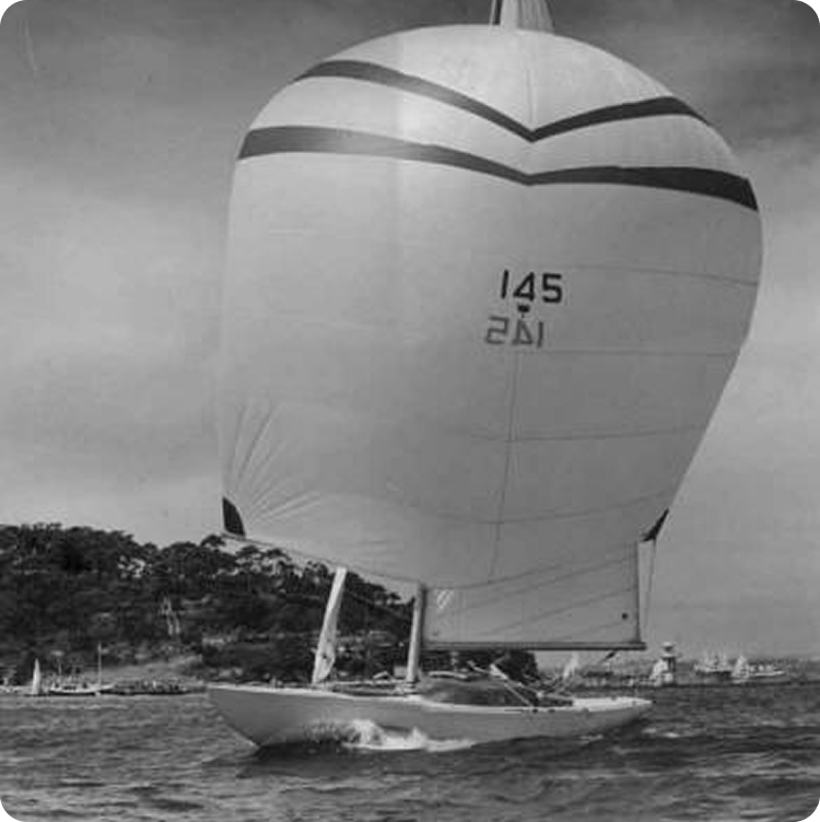 A Schärenkreuzer sailboat with the number 145 on its large, rounded sail glides across the water. Trees and several other Classic Sailing Yachts are visible in the distance under a cloudy sky. The image is in black and white.