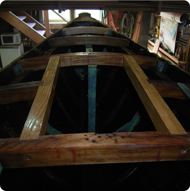 A Schärenkreuzer wooden boat under construction inside a workshop, with frames and ribs exposed, captures the timeless craftsmanship of Classic Sailing Yachts amid tools and shelves in the background.