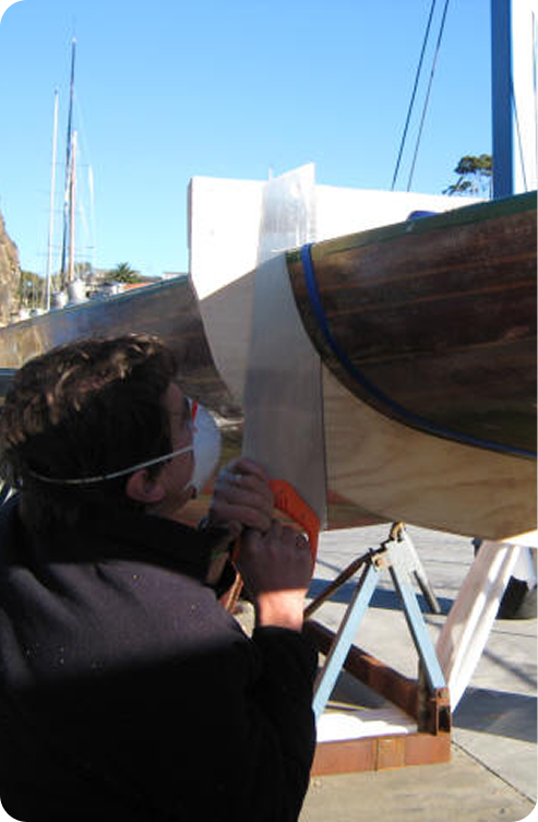 A person wearing a mask and glasses applies fibreglass to the side of a wooden Skerry Cruiser, which is supported on a stand outdoors under a clear blue sky.