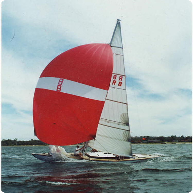 A classic sailing yacht with a large red and white spinnaker sails on a body of water under a partly cloudy sky, with trees and buildings visible in the distant background.
