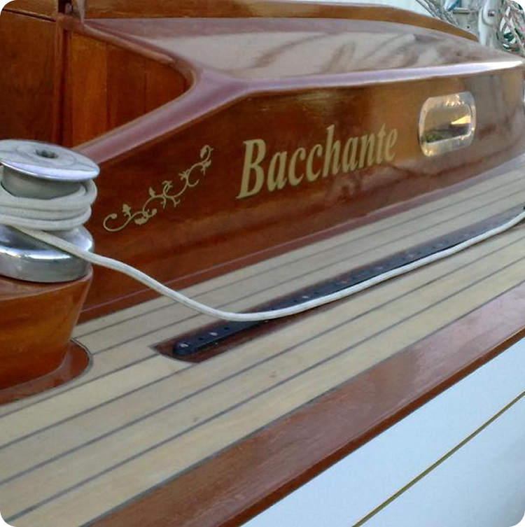 Close-up of a polished wooden boat deck with the name Bacchante painted in gold, showcasing decorative designs and boat equipment with rope—a stunning example of Classic Sailing Yachts craftsmanship.