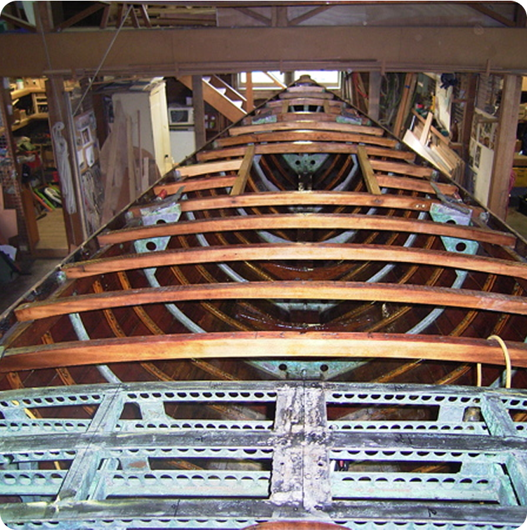A wooden boat, reminiscent of Classic Sailing Yachts or Schärenkreuzer, is under construction in a workshop, revealing its rib structure and framework with metal supports, surrounded by tools and materials.