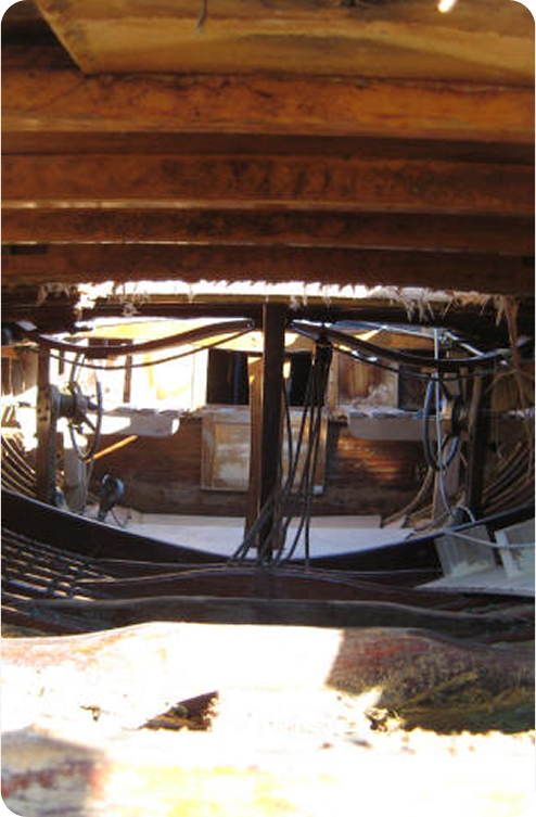 View of the wooden interior of a Schärenkreuzer under construction or repair, with exposed beams, cables, and sunlight filtering through openings above.