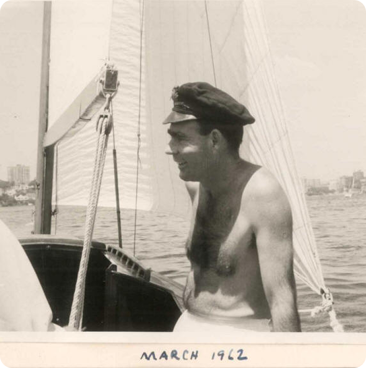A bare-chested man wearing a captain's hat smiles whilst standing on a Skerry Cruiser sailing boat. The background shows water, sails, and distant buildings. The photo is labelled MARCH 1962.