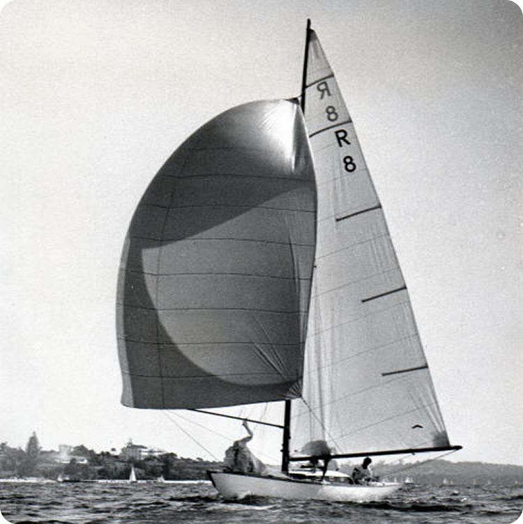 A black and white photo of a Classic Sailing Yacht with a large, billowing sail on the water. Two people are on board, and a shoreline with trees and buildings is visible in the background.