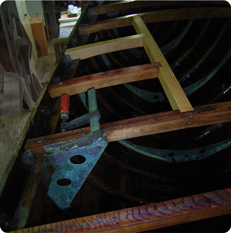 A close-up of a classic sailing yacht under repair, showing its internal ribs, cross beams, and a large metal clamp holding two planks together. Some wood appears new, contrasting with the older, darker timber.
