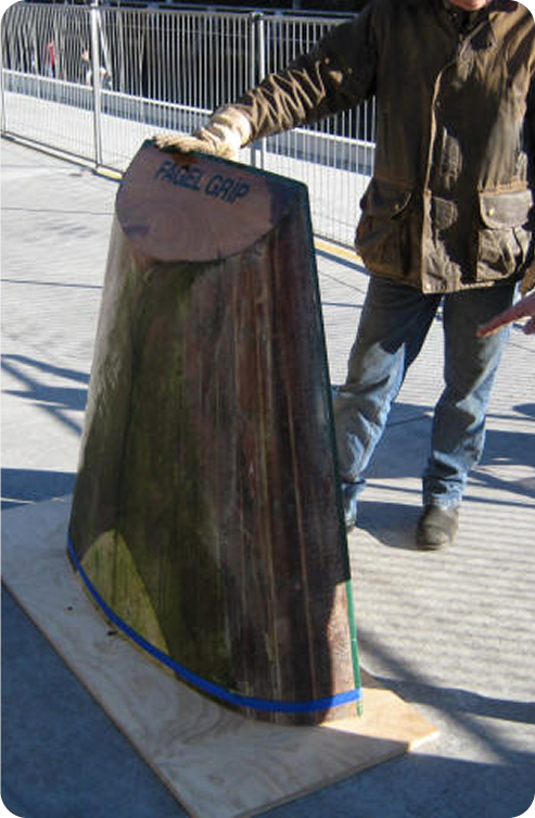 A person stands next to and touches a large, vertically displayed piece of curved glass labelled FACE GRIP, mounted on a wooden base outdoors near a metal fence—reminiscent of the sleek design seen in classic Schärenkreuzer or Square Metre Yachts.