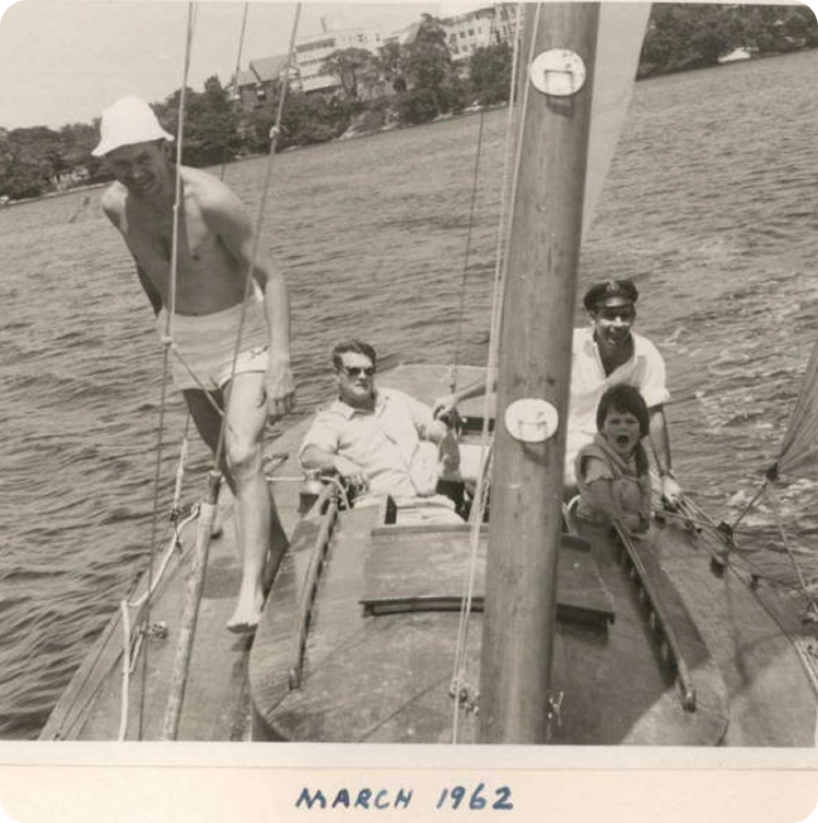 Four people—three adults and one child—enjoy a day on a classic Skerry Cruiser sailboat. One man stands in swimming trunks and a hat, while the others sit or recline. Labelled “March 1962,” trees and buildings line the shore in the background.