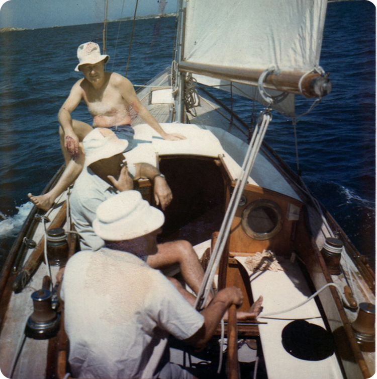 Three men in light-coloured hats relax on a sailboat under sunny skies, with deep blue water surrounding them. Enjoying the timeless elegance of Classic Sailing Yachts, one man sits shirtless at the bow while the others steer near the stern.