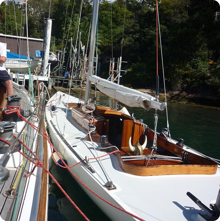 A white Square Metre Yacht moored at a marina with its sail furled, surrounded by calm water and greenery in the background. Red and blue ropes add a dash of colour to this classic sailing yacht’s deck.