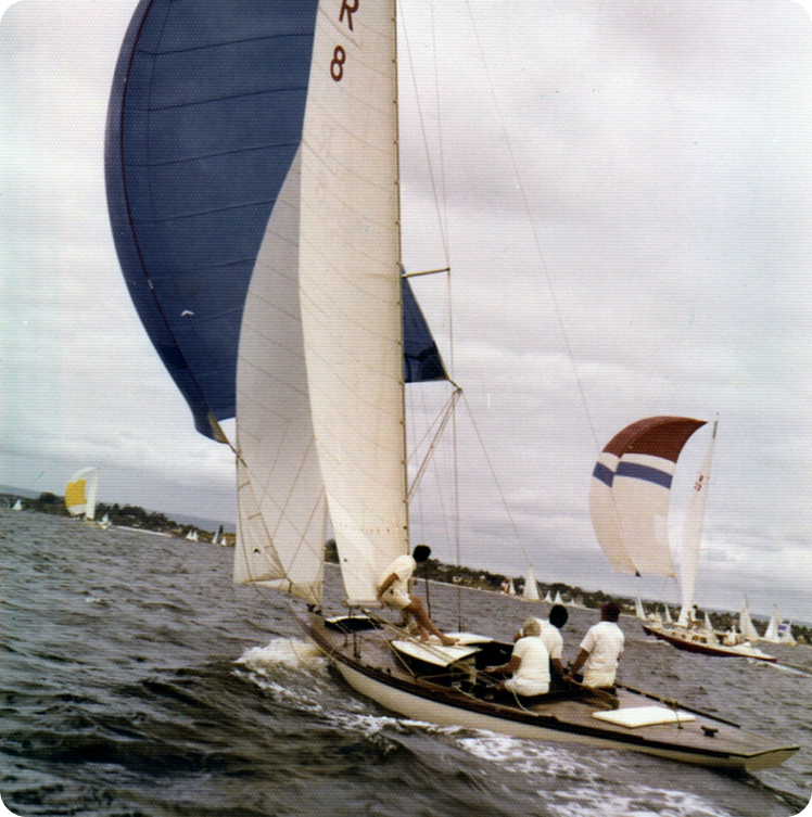 A group of people sail a boat with white sails and a large blue spinnaker on choppy water, with other Square Metre Yachts in the background under a cloudy sky.