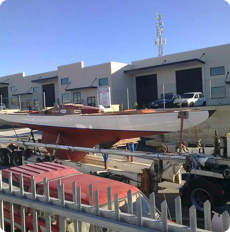 A white and red Schärenkreuzer sailboat sits on a flatbed trailer, parked in an industrial area with modern warehouse buildings, cars, and a blue sky in the background. A metal fence is visible in the foreground.