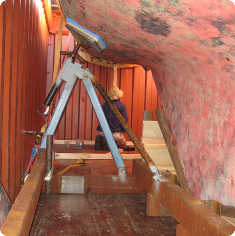 A person sits on the floor inside a shipping container, working near the keel of a weathered pink Skerry Cruiser supported by braces and stands.