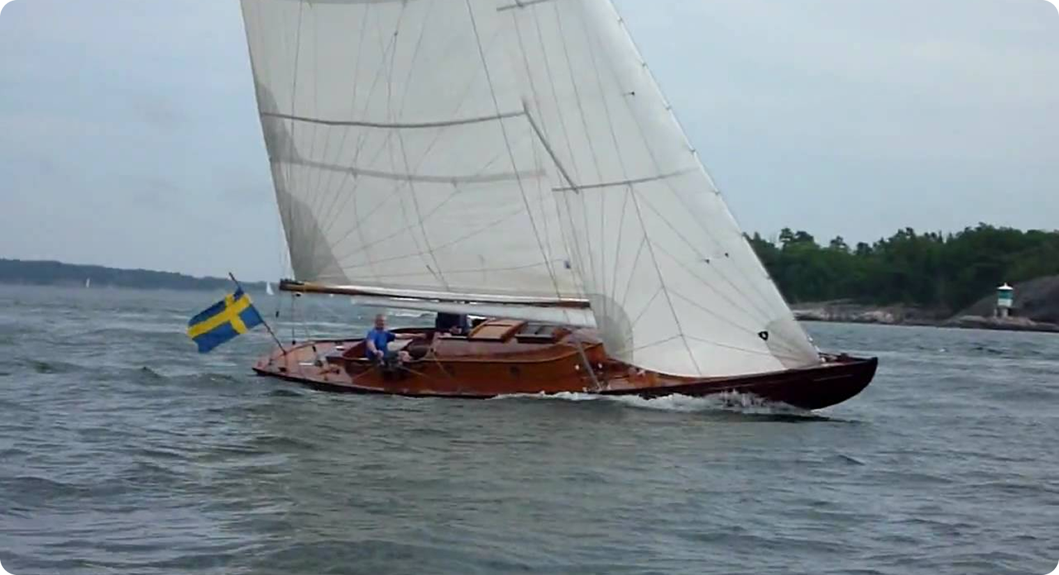 A classic Skerry Cruiser with a Swedish flag sails on choppy waters near a forested shoreline under a cloudy sky. Two people are visible aboard, and the sail is fully extended.