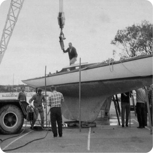 A black and white photo shows several people working together to hoist a Schärenkreuzer sailboat with a crane. One person stands on the boat’s deck, guiding the process, while others stand nearby, securing the Skerry Cruiser and crane.
