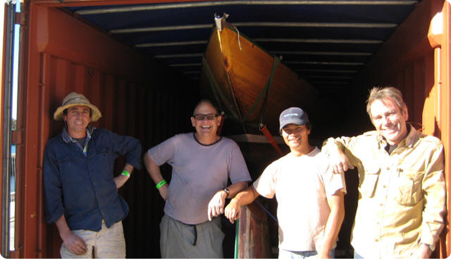 Four men stand smiling inside a shipping container with a yellow boat behind them, likely a Skerry Cruiser or Square Metre Yacht. They are casually dressed in sunhats, T-shirts, and button-downs as sunlight streams into the space.