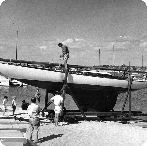 A group of people stands on a sandy shore near a Schärenkreuzer sailboat resting on a wooden support structure, with one person working on the boat and others observing. Additional Skerry Cruisers and water are visible in the background.