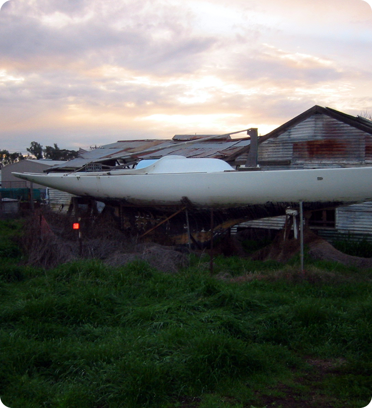 A large, white Skerry Cruiser sailboat sits on supports in overgrown grass beside an old, weathered, tin-roofed shed at sunset, with a cloudy sky in the background.