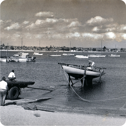 A black-and-white photo shows two men preparing to launch or retrieve a Classic Sailing Yacht from a calm bay using a trailer. Several boats are moored in the water, and houses line the distant shore under a partly cloudy sky.