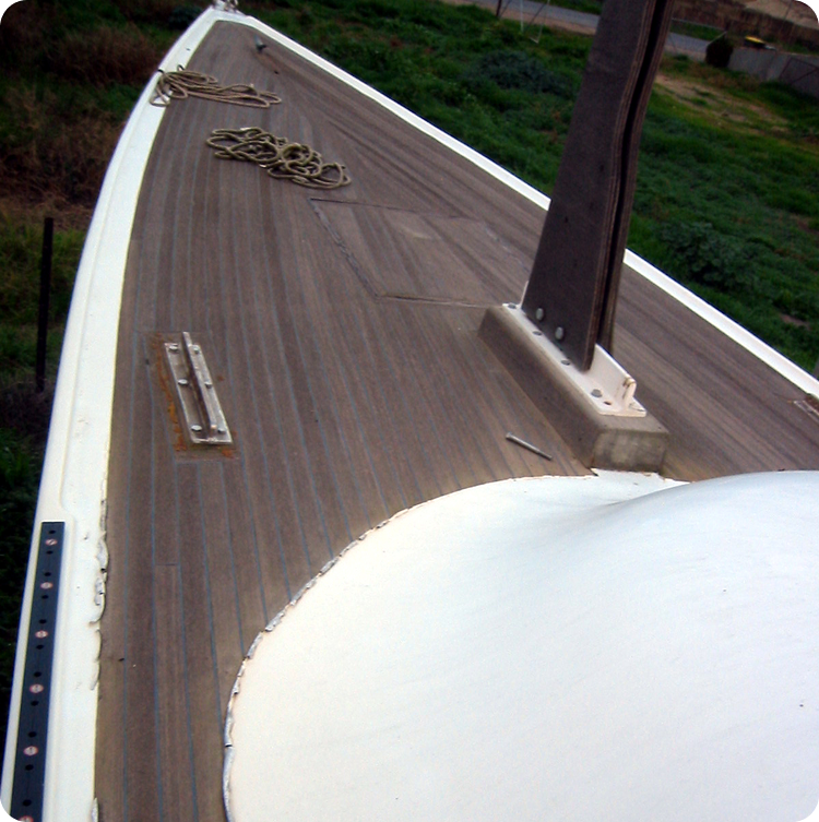 View of a Skerry Cruiser’s wooden deck with a coiled rope and a mast rising from the centre. The weathered Classic Sailing Yacht rests against greenery in the background, evoking timeless maritime elegance.
