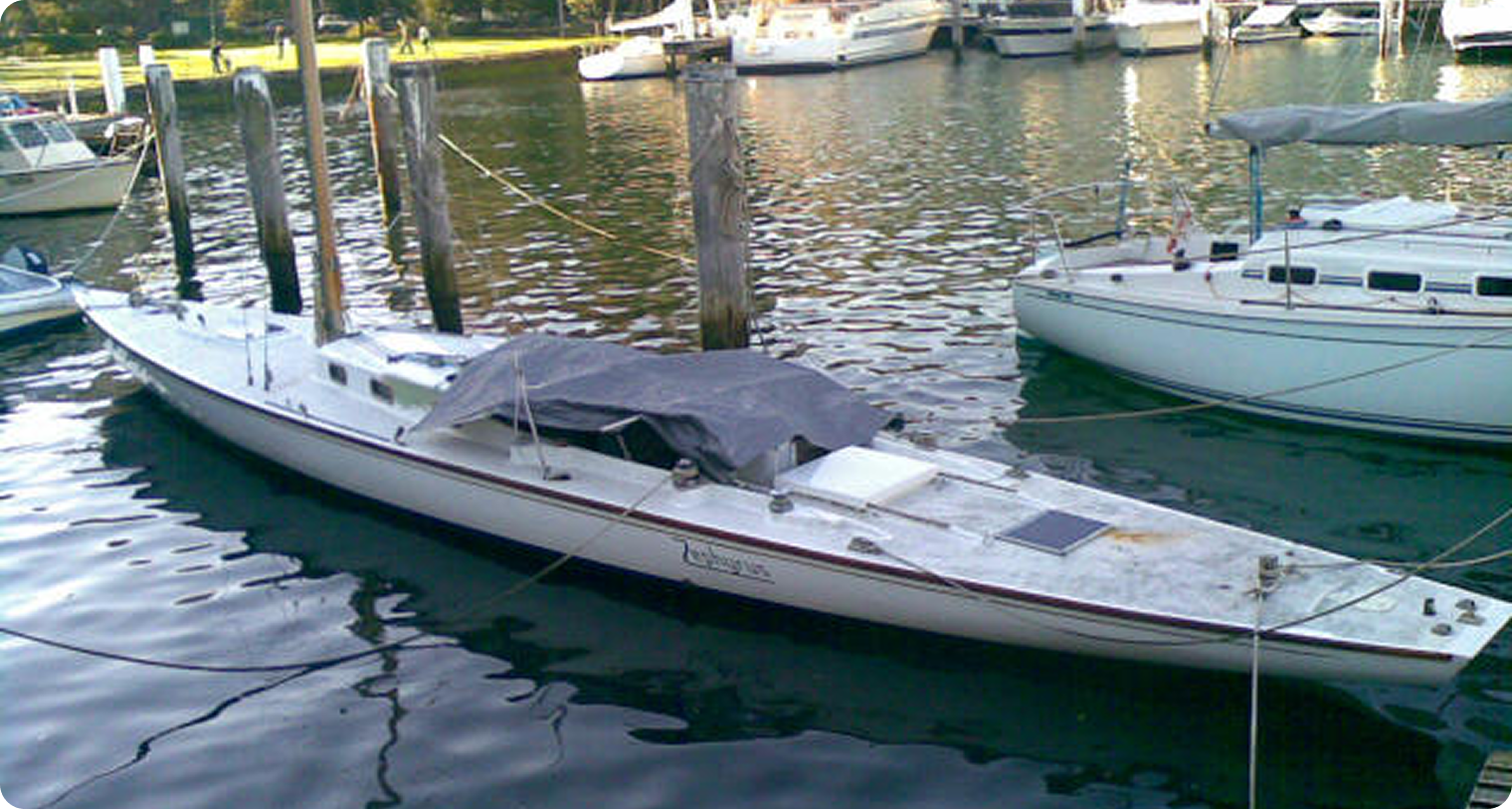 A long, narrow white Classic Sailing Yacht covered with a grey tarpaulin is docked at a marina, moored alongside wooden posts. Other boats and yachts are visible in the background on calm water.