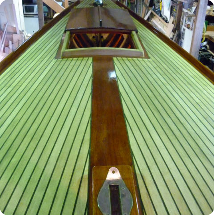 A close-up view of a Schärenkreuzer boat deck with light green planks and polished wooden trim, photographed indoors in a workshop with tools and materials visible in the background.