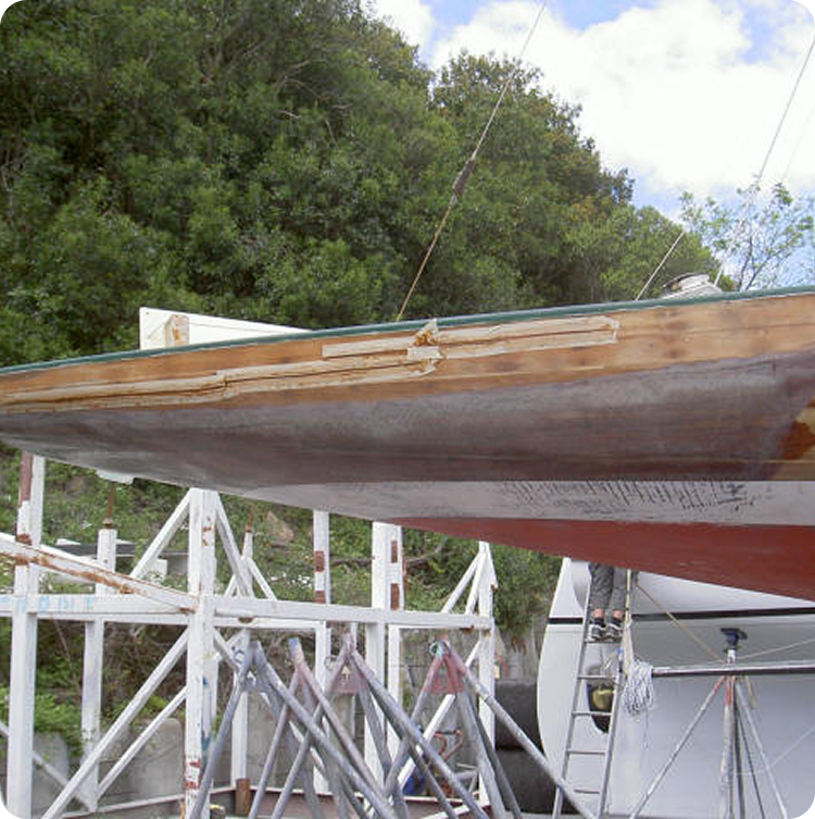 A close-up of a Skerry Cruisers boat hull undergoing repair, supported by metal stands. The hull has visible patches of fresh woodwork and filler, with trees and a cloudy sky in the background.