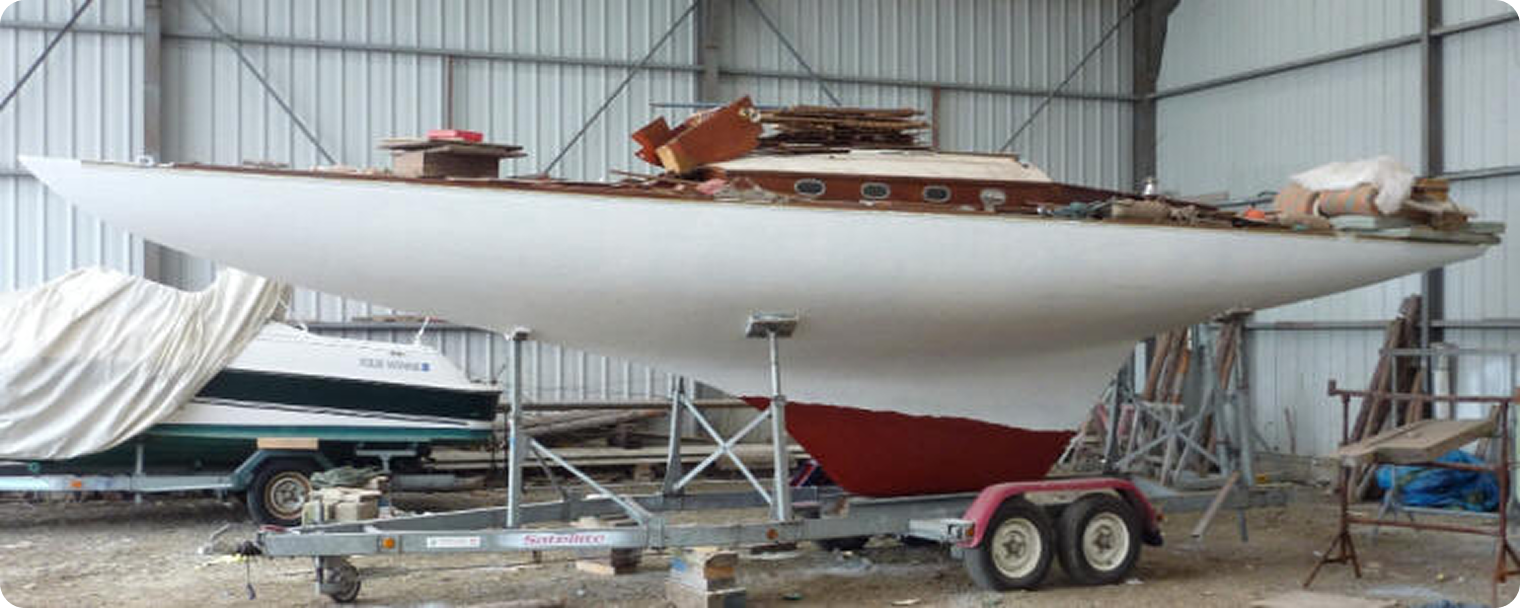 A white sailboat with a red keel, likely one of the classic Square Metre Yachts, sits on a trailer inside a large metal shed; various materials and wooden planks are stacked on its deck, and another covered boat is nearby.