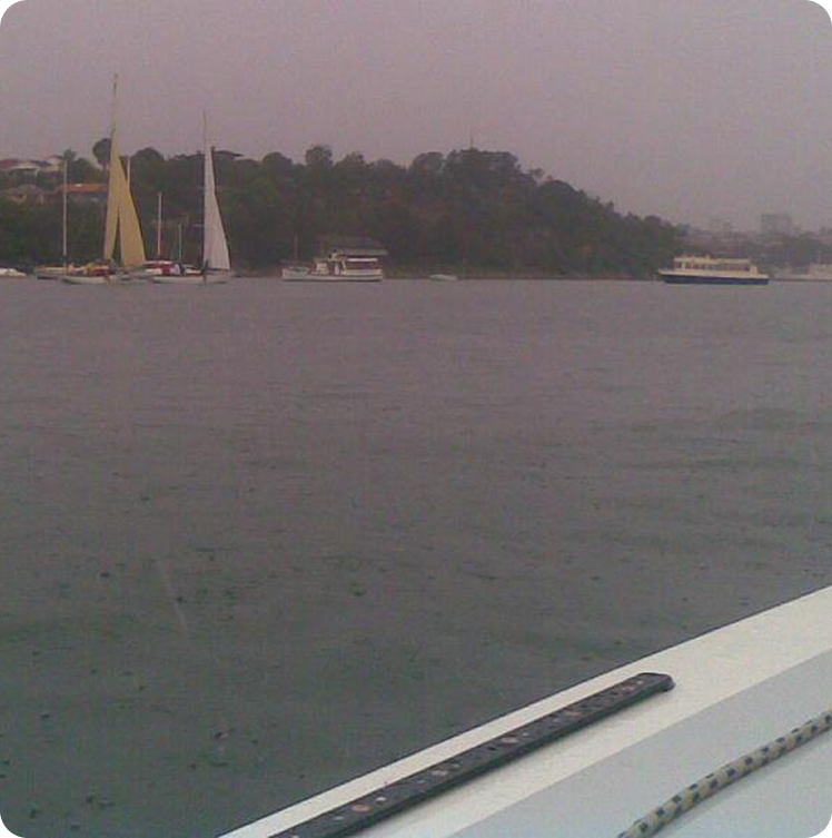 View from a boat on a cloudy day, showing a wide river or bay with Classic Sailing Yachts and ferries in the distance near a tree-lined shore.