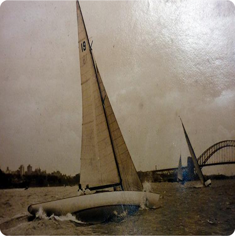 A vintage photo shows two Classic Sailing Yachts racing on choppy water, with one prominently heeling to the side. In the background, the Sydney Harbour Bridge arches under a cloudy sky.