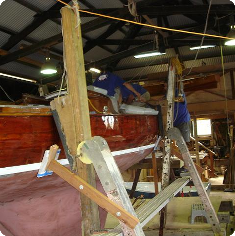 Two people in a workshop are working on the side of a polished brown Schärenkreuzer, using ladders and scaffolding. Tools and building materials surround the elegant Square Metre Yacht as they restore its classic beauty.