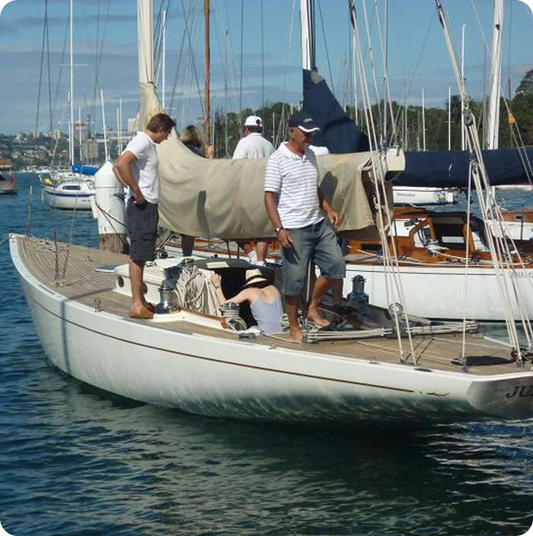 Two men stand on the deck of a classic sailing yacht moored in a marina, with other boats and people in the background on a sunny day.