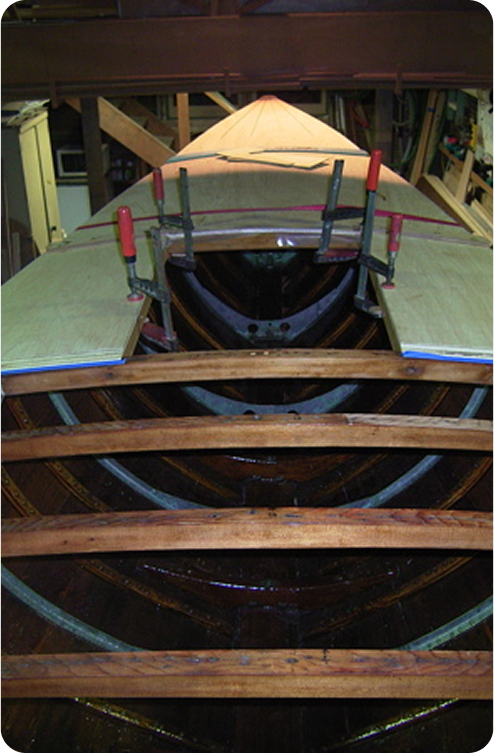 Wooden Skerry Cruiser under construction in a workshop, showing exposed framework and ribs. Several clamps hold pieces of wood in place on the deck, with woodworking tools and materials visible in the background.